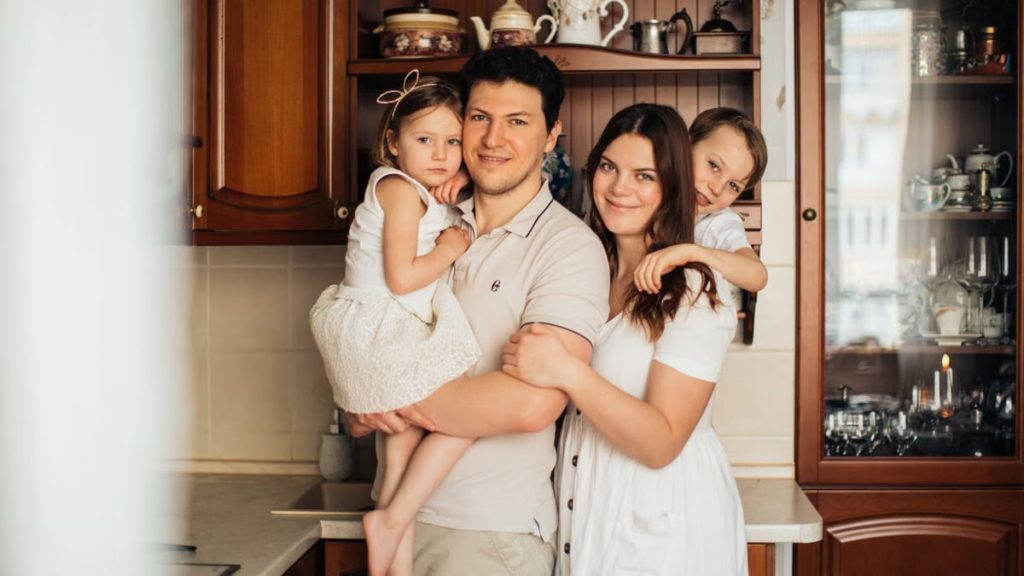 Satisfied man with daughter and woman with son embracing embracing each other while standing against wooden cupboard in kitchen and looking at camera